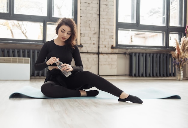 Fit brunette vrouw met fles water zit op mat in yogastudio