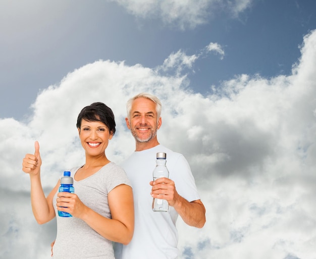 Fit couple with water bottles gesturing thumbs up against blue sky with clouds