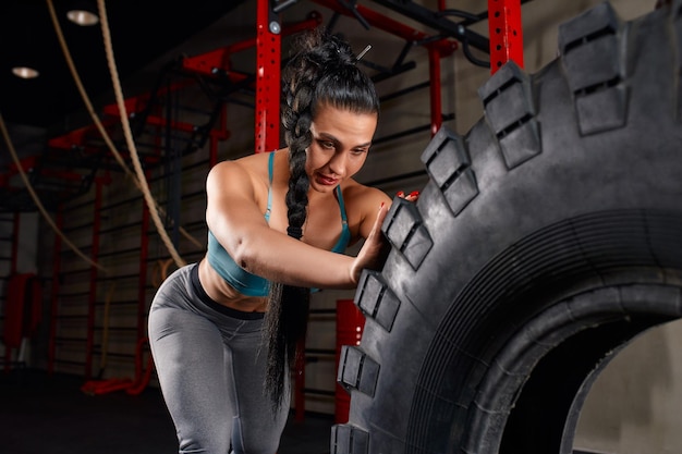 Atleta femminile in forma che si allena con un enorme pneumatico girando e capovolgendo in palestra donna che si esercita con un grosso pneumatico