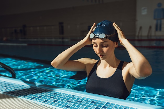 fit female swimmer in cap and swimming goggles preparing for training in sunlit indoor pool