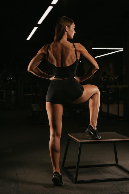 A fit girl in a black suit is posing with her back in a gym. A sporty girl is stretching after a workout.