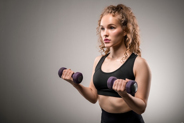 Fit girl with blond curly hair holding dumbbells with her arms bent in elbows while exercising in gym on grey