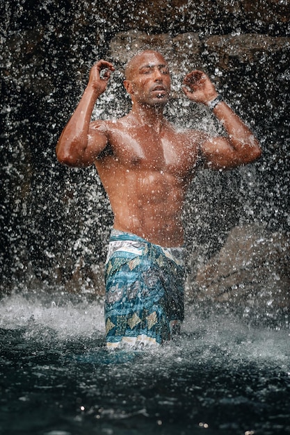 Подтянутый и красивый топлесс арабский мужчина под водопадом, огромные камни и вода, брызги и блики; концепция естественной красоты.