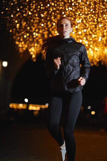 Fit jonge roodharige vrouw in sportkleding die langs de weg in de stad loopt 's nachts verlicht door licht...