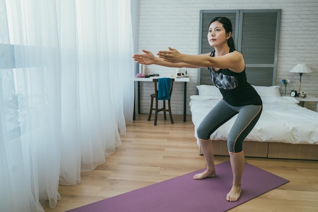 fit Korean female is doing squat on the exercise mat with parallel arms by the window on sunny morning in the bedroom during home workout.