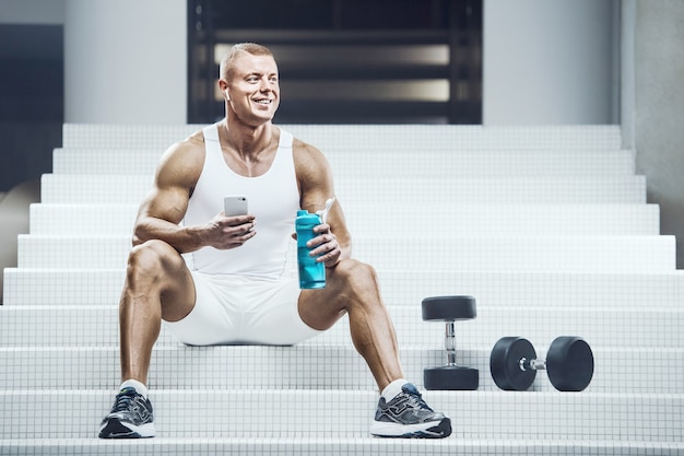 Fit man sitting down in white stairs with a blue shaker and some weights