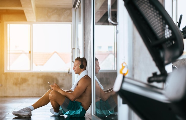 Photo fit man using headphone and listening to music while sitting after workout at the gym