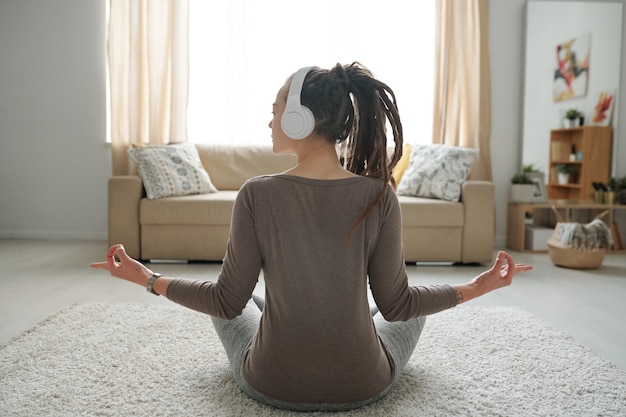 Fit meisje in activewear luisteren naar ontspannende muziek in koptelefoon zittend in de pose van lotus thuis