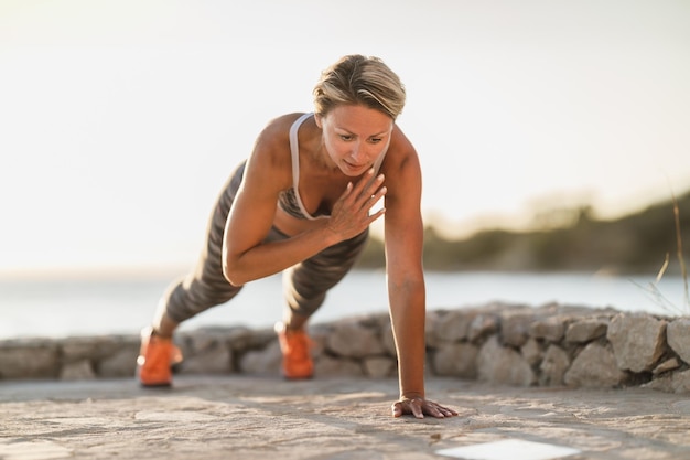 Fit middle age woman doing plank or push-up exercises during hard training near the sea beach.