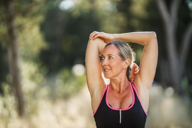 Fit middle age woman doing stretching exercises during training in pine forest near sea shore.