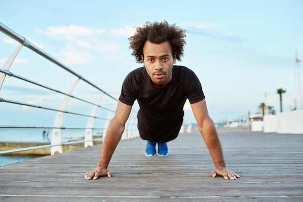 Fit and motivated Afro-American sportsman standing in plank position, stretching on wooden platfom. Dark-skinned sportsman with bushy hair doing yoga at seaside.