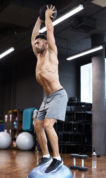 Fit and muscular man exercising with medicine ball on gymnastic hemisphere bosu ball in gym.