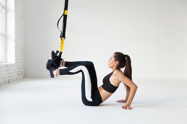 Fit pretty young woman doing fly yoga stretching exercises with trx fitness straps  in fitness training white gym loft classroom. Sport healthy lifestyle concept. Copy space.