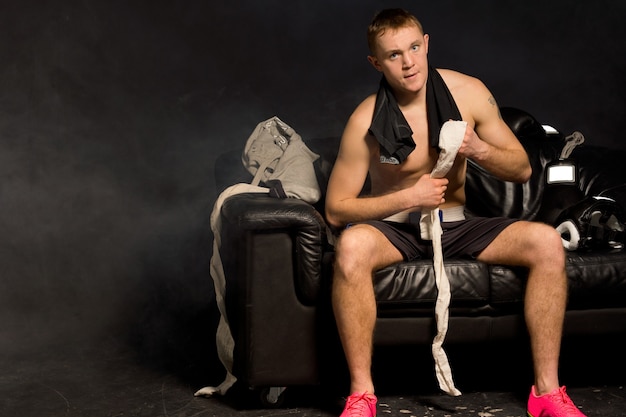 Серьезный молодой боксер с мускулистым телосложением сидит, перевязав руки, на черном кожаном диване.