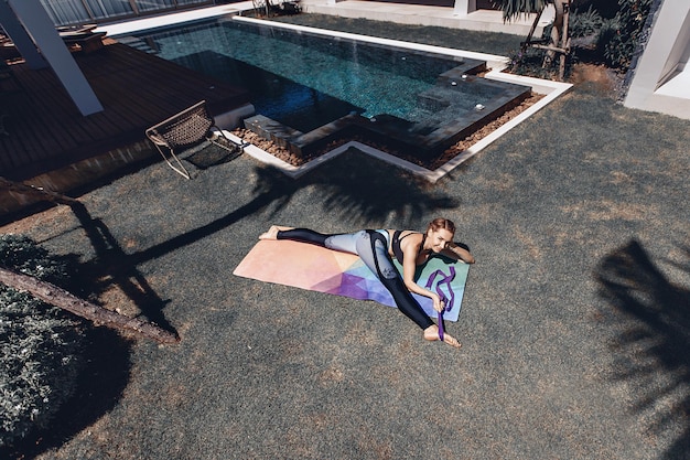 Fit sexual skinny model doing yoga and developing her flexibility while lying on a yoga mat on the outdoor sports field. Summer day. Sport time concept