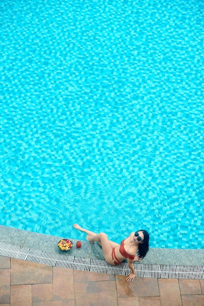 Fit slim young woman in bikini and sunglasses sitting on edge of swimming pool of spa resort