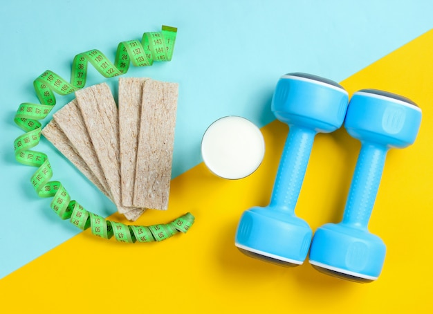 Fit slimming concept. Dumbbells, ruler, glass of kefir, diet bread on a blue-yellow table