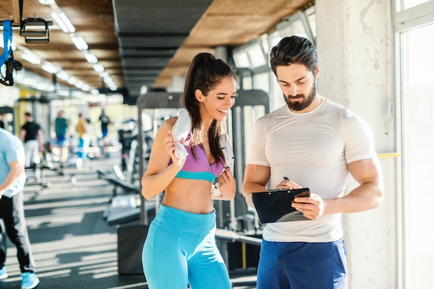 Fit smiling Caucasian woman with towel around neck looking at clipboard. Her personal trainer writing and showing her result of training. Gym interior.