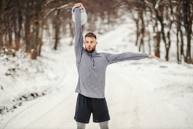 Fit sportman die kettlebell opheft terwijl hij op een besneeuwd pad in het bos staat. Winterfitness, gezond leven, bodybuilding