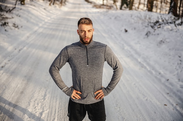 Fit sportman staande op besneeuwde pad in de natuur en hand in hand op de heupen. Wintersport, winnaar, prestatie