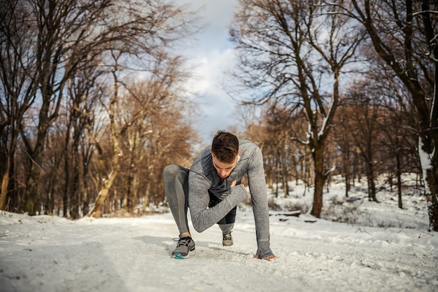 눈 덮인 겨울 날 자연 속에서 무릎을 꿇고 워밍업과 스트레칭 운동을 하는 스포츠맨. 겨울 피트니스, 건강한 라이프 스타일