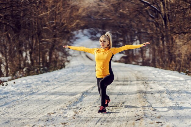 Fit sportswoman doing lunges on snowy path at sunny winter day. Warm up exercises, winter fitness, healthy habits