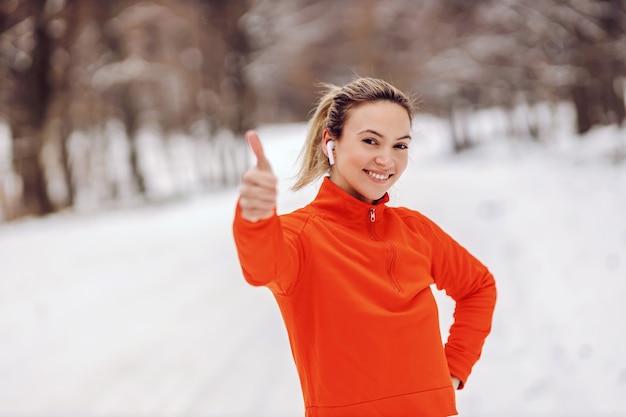 Sportiva adatta in piedi nella natura il giorno nevoso e mostrando i pollici. approvazione, stile di vita sano, clima freddo, neve, fitness invernale