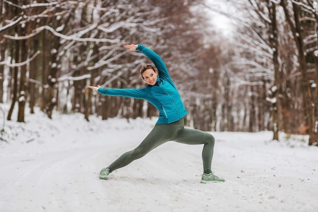 Fit sportswoman in warm outfit doing yoga exercises in snowy forrest. Fitness outdoors, snow, cold, winter yoga