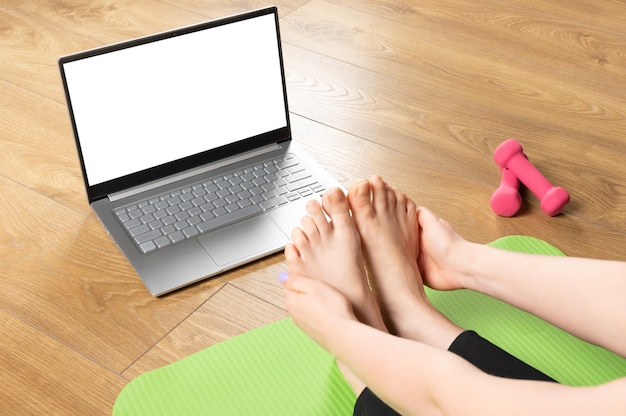 Fit sporty healthy woman sitting on mat in Paschimottanasana pose, doing breathing exercises, watching online yoga class on mockup laptop