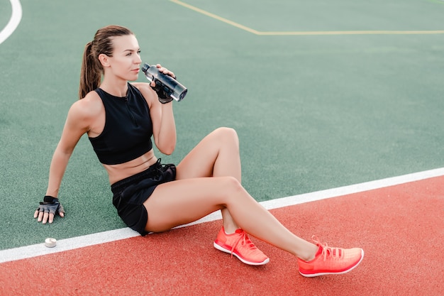 Fit sporty woman drinking water from the bottle on stadium during fitness workout