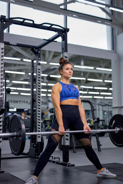 強い白人女性をスポーティーな服のトップに合わせ、バーベルでトレーニングして、引っ張ったり、しゃがんだり、ジムでフィットネスをしたり、足を離して立ったりします。スポーツ、トレーニング、ボディービルの概念。