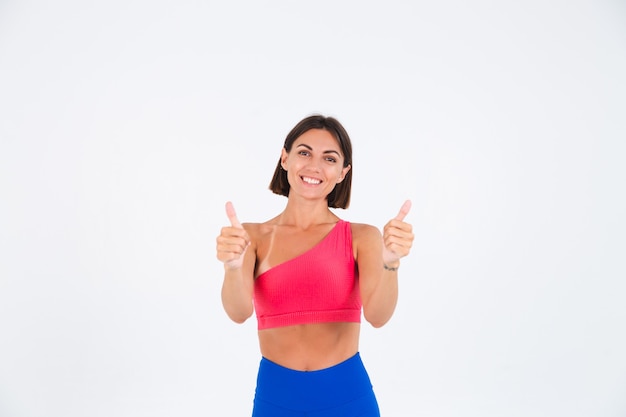 Fit tanned sporty woman with abs, fitness curves, wearing top and blue leggings on white shows thumbs up with smile