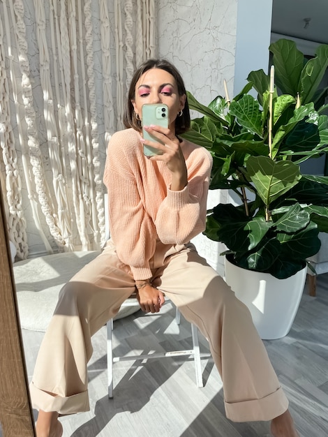 Fit tanned woman in peach sweater and classic beige trousers at home taking photo selfie