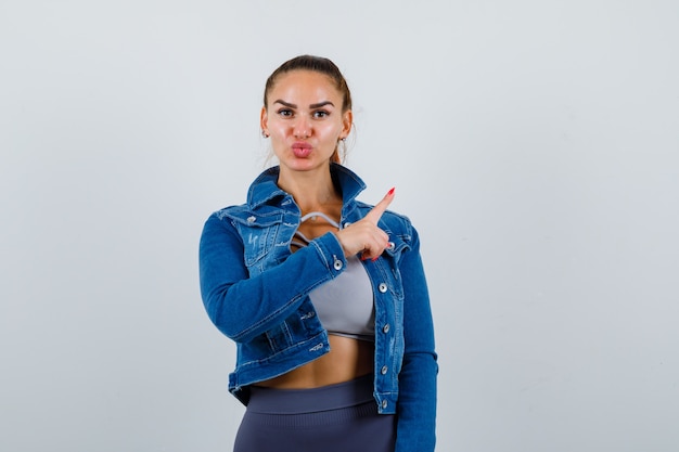 Fit vrouw die met wijsvinger naar rechts wijst, kusjes stuurt in crop top, spijkerjasje, legging en er schattig uitziet. vooraanzicht.