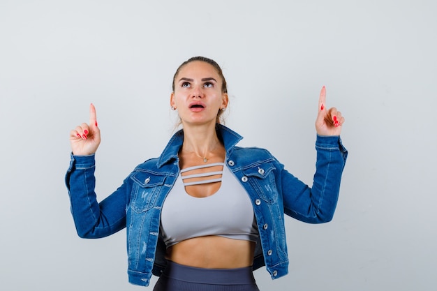 Fit vrouw die met wijsvingers omhoog wijst in crop top, spijkerjasje, legging en verbaasd kijkt. vooraanzicht.