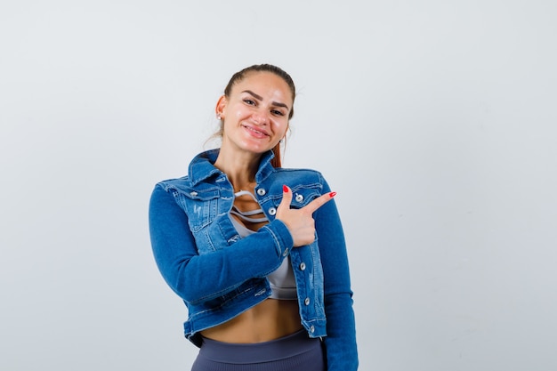 Fit vrouw die naar rechts wijst met wijsvingers in crop top, jeansjack, legging en er gelukkig uitziet, vooraanzicht.