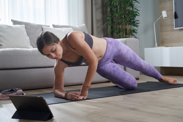 Fit vrouw die yoga beoefent op haar mat met behulp van een online trainingsprogramma op haar tablet