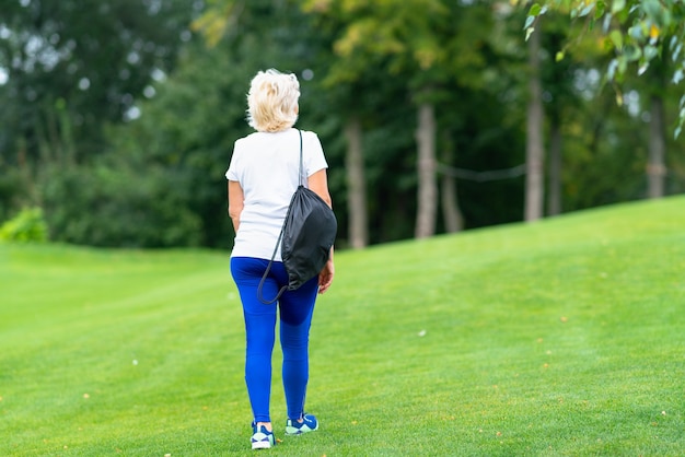 Fit woman carrying a sports bag over her shoulder walking away over the green grass in a park towards distant trees