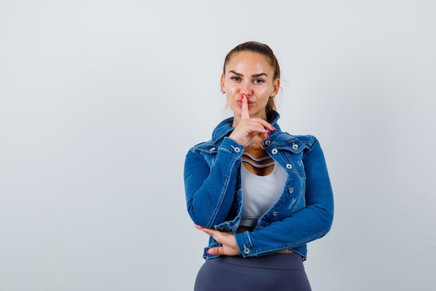 Fit woman in crop top, jean jacket, leggings showing silence gesture and looking serious , front view.