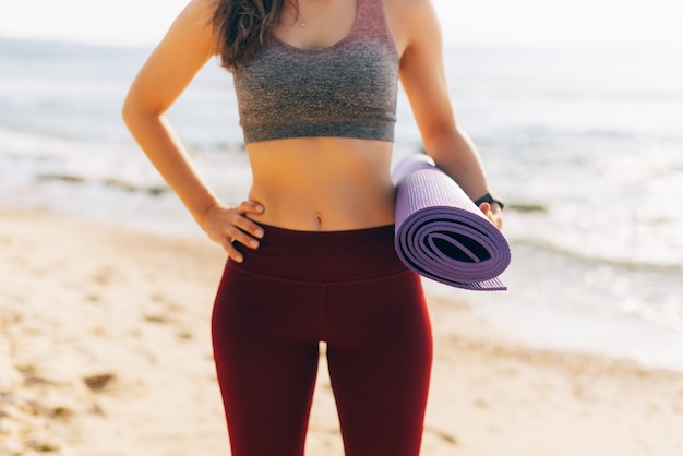 Fit woman is holding a violet mat while standing on the beach.