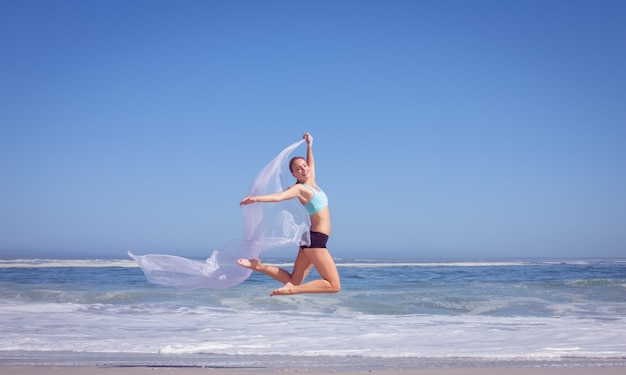 Fit woman jumping gracefully on the beach with scarf