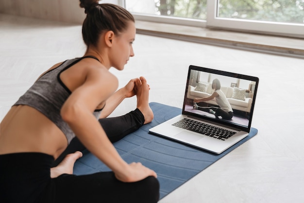 Montare la donna in abiti sportivi che fa yoga sulla stuoia di esercizio a casa utilizzando laptop guardando tutorial. l'allenatore femminile ha una lezione di yoga virtuale sul computer