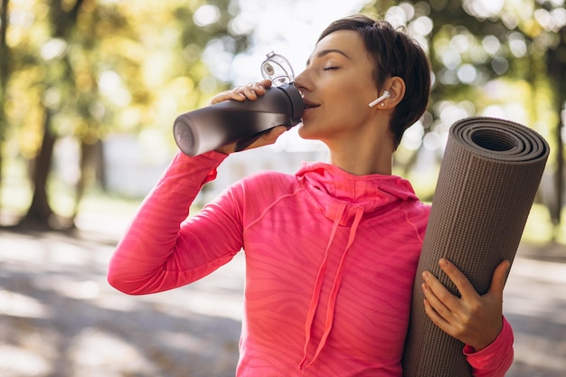 Fit woman srinking water in park and holding yoga mat