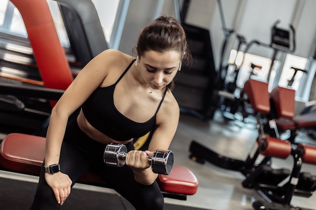 Fit woman training biceps doing concentrated lifting dumbbell for biceps while sitting on a bench in the gym
