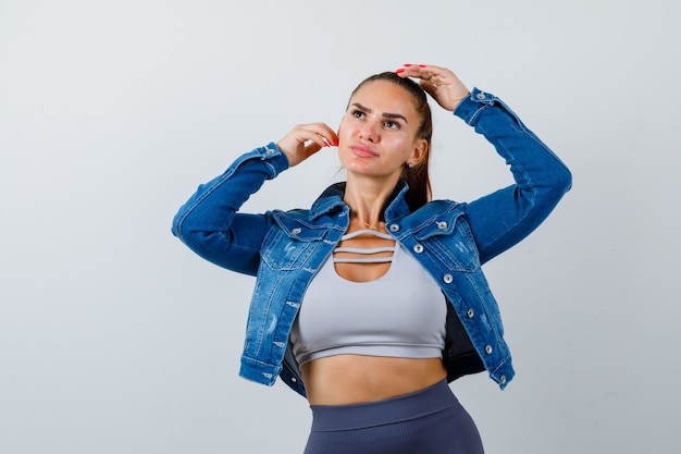 Donna in forma con una mano sulla testa, un'altra mano sulla guancia mentre posa in top corto, giacca di jeans, leggings e sembra sicura. vista frontale.