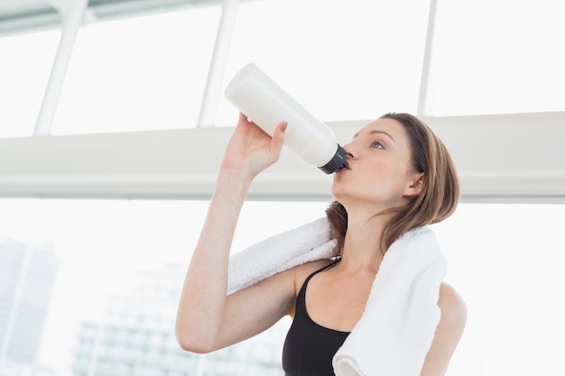 Fit woman with towel around neck drinking water in fitness studio