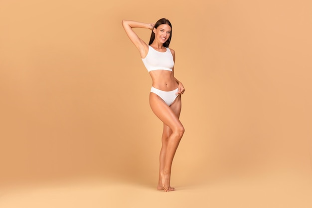 Подтянутая юная леди с гладкой кожей и идеальной формой тела позирует в белом нижнем белье на бежевом фоне в полный рост