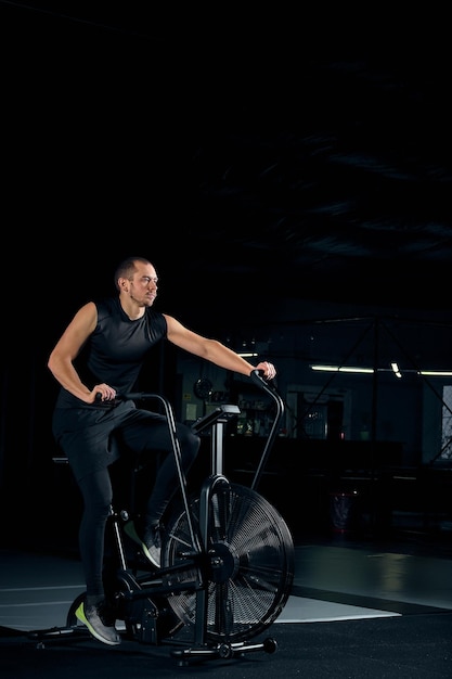 ジムでエアロバイクを使用して若い男にフィットします。クロストレーニングジムで有酸素運動にエアバイクを使用しているフィットネス男性。