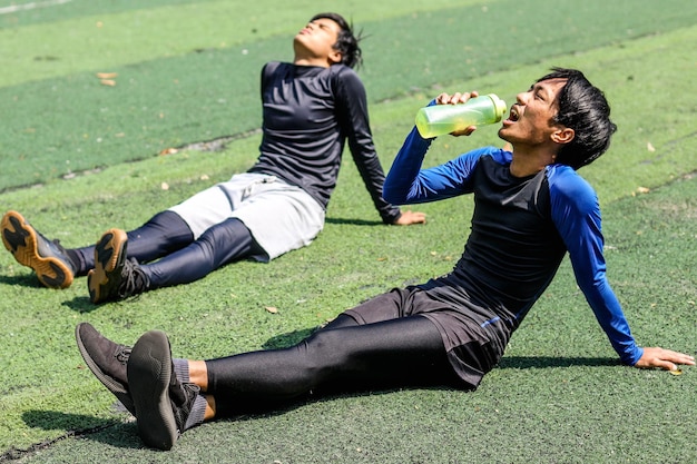 Fit young men taking rest from running by drinking mineral water from tumbler Two friends taking a