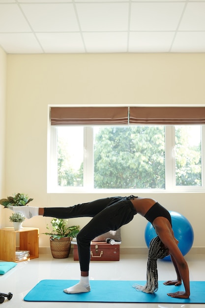 Fit young woman standing in wheel pose on yoga mat and raising one leg